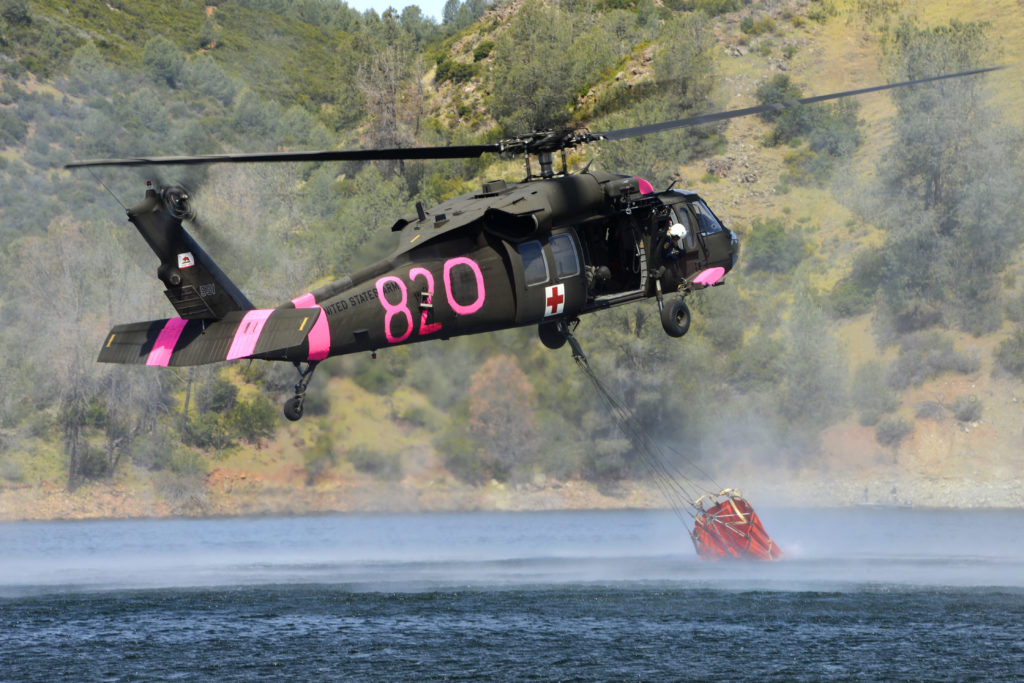 Seis UH-60 Black Hawk “bombeiros” para Portugal. Um helicóptero UH-60 Black Hawk enche um bambi bucket com água durante o treinamento de combate a incêndios com  helicópteros militares interagências da Califórnia na academia de combate a incêndios do estado, perto de Pardee Dam em Ione, Califórnia (US DoD).